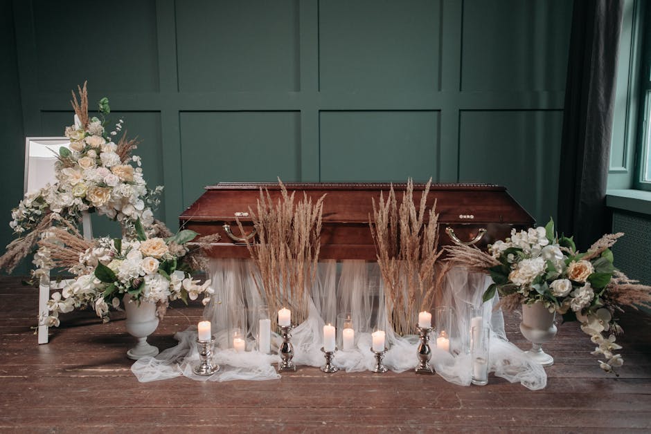 A carefully arranged floral tribute for a funeral, featuring two large white urns filled with cream and blush roses, white hydrangeas, and soft pink carnations, accented with pampas grass and lush gre