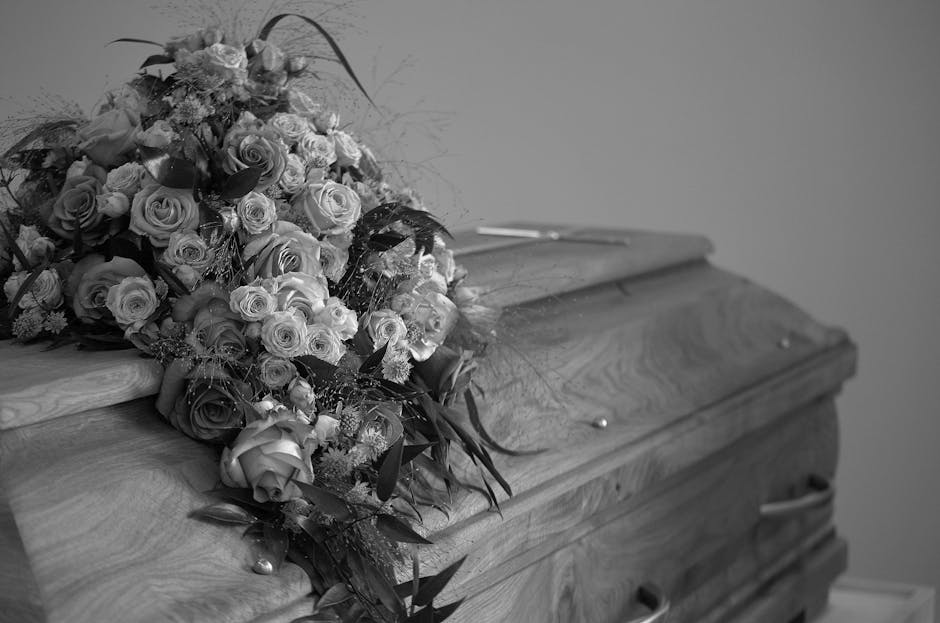 A bouquet of fresh flowers placed on top of a wooden casket. The arrangement features a mix of roses, some fully bloomed and others in bud form, with various shaded petals, predominantly light-colored