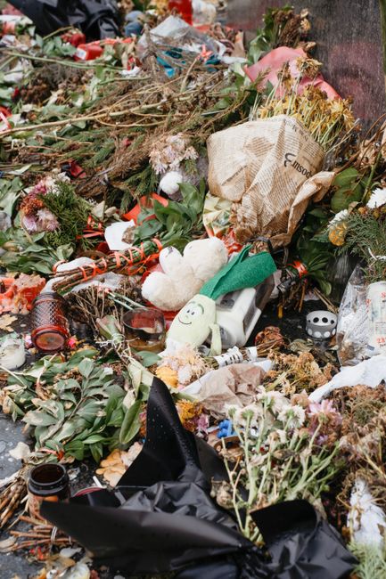 A chaotic scene of discarded flowers and floral packaging, including wilted pink roses, dried foliage, and various crumpled wrapping papers. Among the debris, there are broken stems, a wrapped bouquet