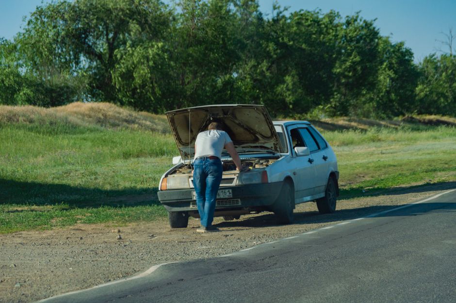 A woman with long hair wearing a white top and blue jeans is standing by a light blue car with its hood open, inspecting the engine. The car is parked on the side of a rural road with a grassy verge a