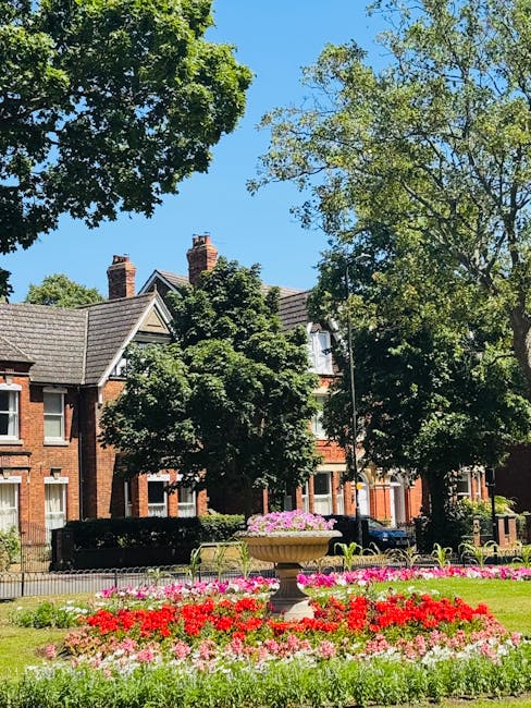 A lush outdoor garden scene featuring a large stone planter filled with pink and purple flowers positioned on a well-maintained lawn. Surrounding the planter are vibrant red, pink, and white blooming