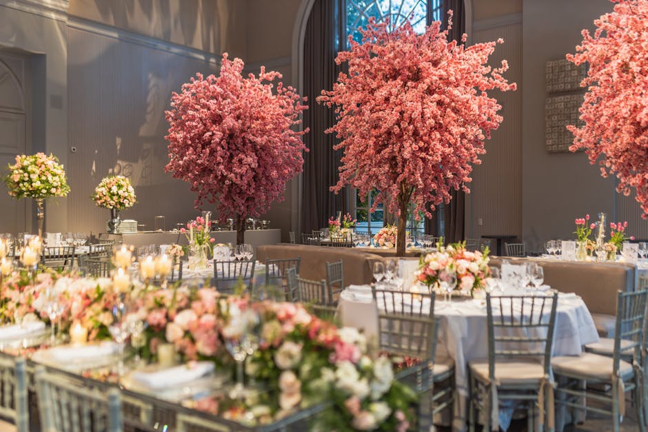 An elegant indoor event space decorated with tall, lush pink cherry blossom trees serving as striking centerpieces. Surrounding the trees are round tables adorned with floral arrangements featuring so