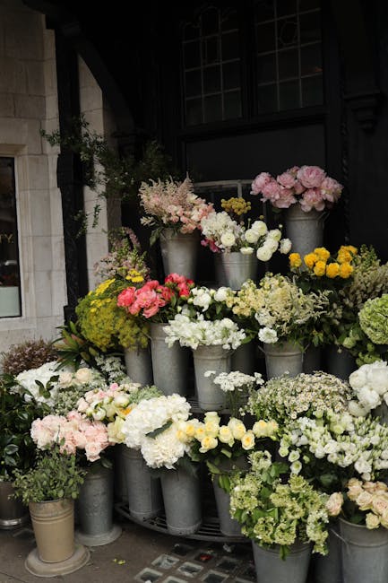 A display of fresh flowers arranged in metal buckets placed on a rounded stand outside a building with dark window frames and a stone wall. The floral composition includes pink, white, yellow, and gre